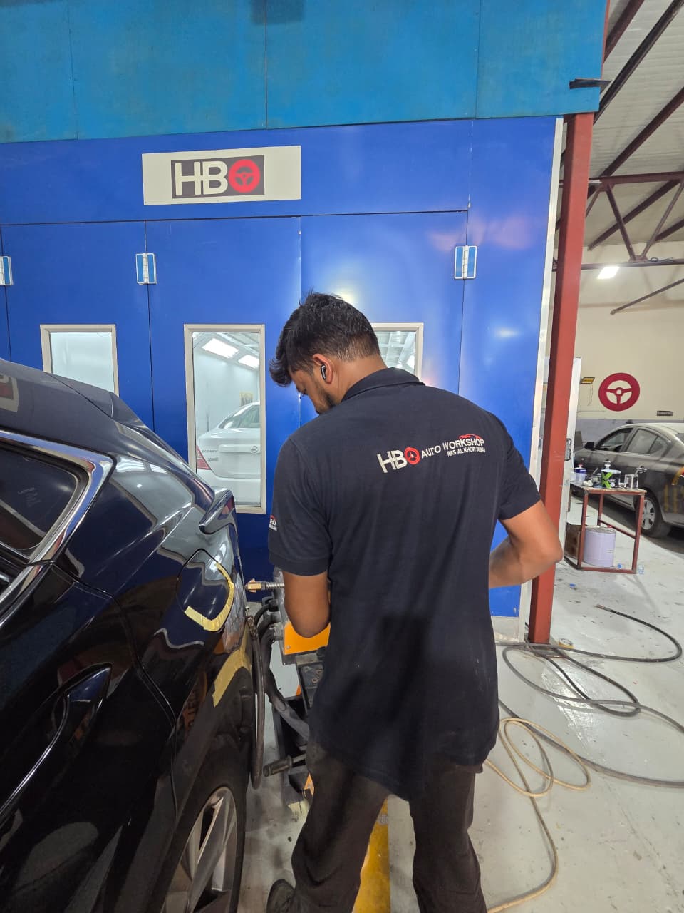 Technician in HBO uniform carrying out paint work on a vehicle panel beside the workshop's blue paint booth